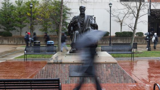 a student walking in the rain with a black umbrella past the patterson office tower statue.